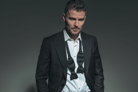 handsome young businessman with undone tie in tuxedo posing in a fashion light with hands in pockets...