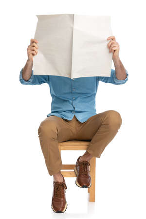 casual man in denim shirt and chino pants with sneakers covering face with newspaper while reading and sitting on wooden chair on white background in studioの写真素材