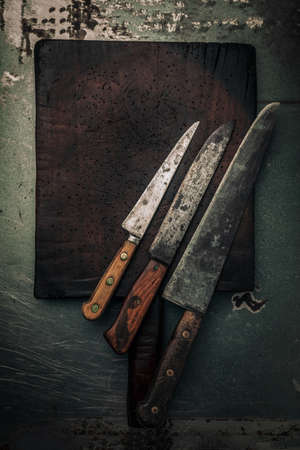 table top picture of set of 3 carbon steel knives on rustic wooden chopping board on rusty metallic background, food and cooking conceptの写真素材