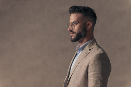 side view of handsome young man in suit with open collar shirt looking to side and posing on beige...