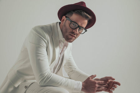 side view of seated fashion businessman in white suit with hat touching palm with finger and posing...