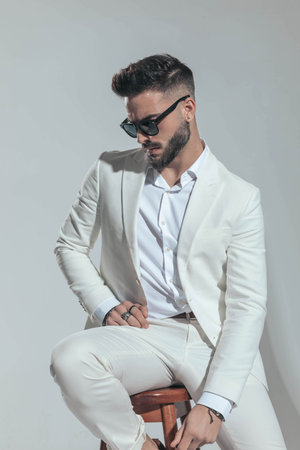 bearded fashion businessman in white suit looking down side and sitting on grey background in studioの写真素材
