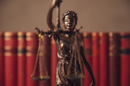 bronze blindfolded goddess representing law statue holding a scale symbolizing impartiality and two broad sources of law in a university room with a background made of booksの写真素材