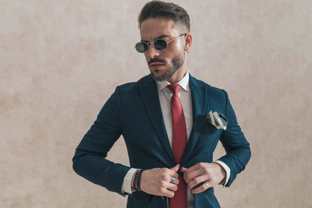 sexy young guy with sunglasses adjusting and opening navy blue suit, wearing red tie and handkerchief, looking to side and posing in studioの写真素材