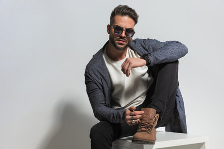 attractive young man with sunglasses holding elbow on knee and posing with leg up while sitting on grey background in studioの写真素材