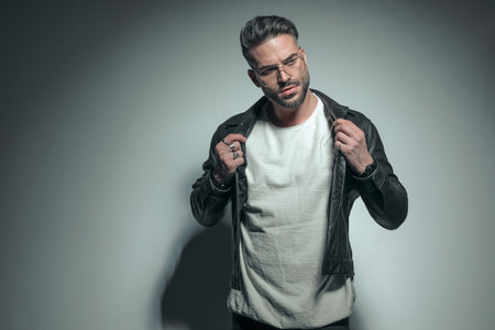 handsome casual man arranging his leather jacket, looking to side and wearing eyeglassesの写真素材