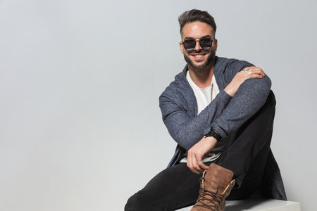 portrait of happy man in hoodie with sunglasses holding elbow on knee and posing while sitting with leg up on grey backgroundの写真素材