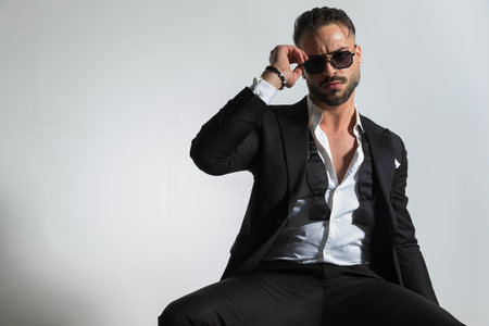 stylish businessman in black tuxedo with untied shirt and bowtie adjusting sunglasses while sitting and posing on grey background in studioの写真素材