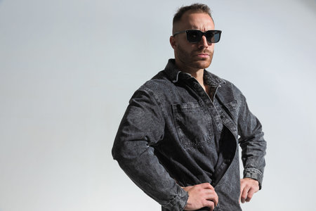 casual young man posing in a funny way while wearing grey denim jacket and looking away on grey...