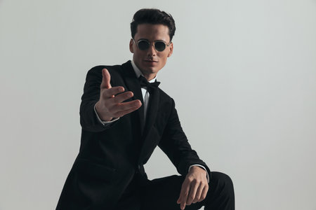 happy young businessman wearing black tuxedo and making an invitation with his hand while posing in front of grey background in studioの写真素材