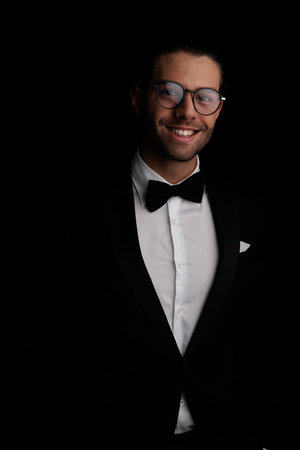 portrait of attractive young groom wearing tuxedo and glasses, being happy and laughing in front of black background in studioの写真素材
