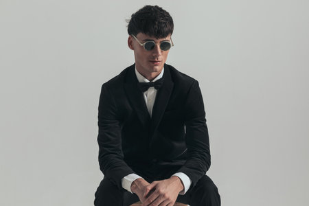 Portrait of attractive businessman rubbing his hands and looking away, sitting on a wooden chair, wearing a black tuxedo and sunglasses, in a fashion poseの写真素材