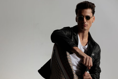 sexy casual guy wearing leather jacket and posing with elbow on knee in front of grey background in...