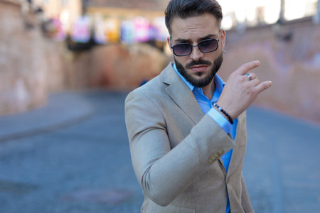 portrait of attractive bearded businessman holding arm in fashion pose and looking down while confidently posing outdoor in an old cityの写真素材