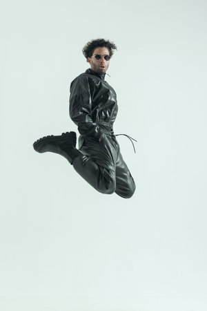 attractive casual man jumping and kicking with hands in pockets, wearing a leather costume in a fashion poseの写真素材
