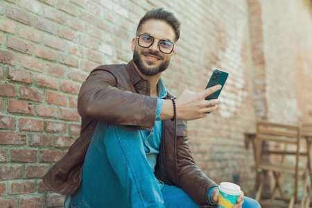 Portrait of handsome casual man having fun with his phone and drink, wearing eyeglasses and leather jacket, outdoor in an old medieval townの写真素材