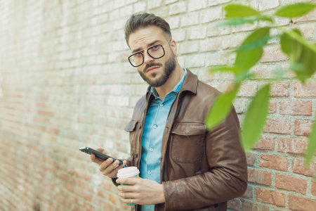 Portrait of attractive casual man texting on phone and holding drink against wall, wearing eyeglasses and leather jacket, outdoor in an old medieval townの写真素材