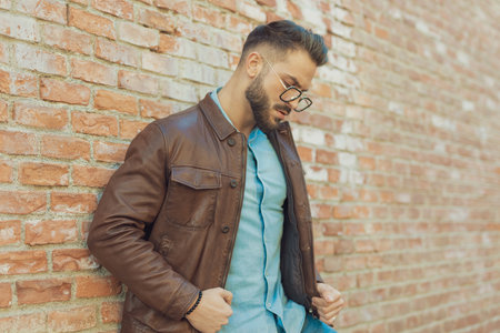 Portrait of sexy casual man leaning on brick wall and arranging his jacket, wearing eyeglasses, outdoor in an old medieval townの写真素材