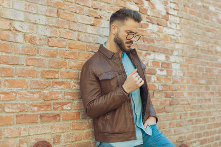 Portrait of attractive casual man leaning on brick wall with hand in pocket, wearing eyeglasses and leather jacket, outdoor in an old medieval townの写真素材