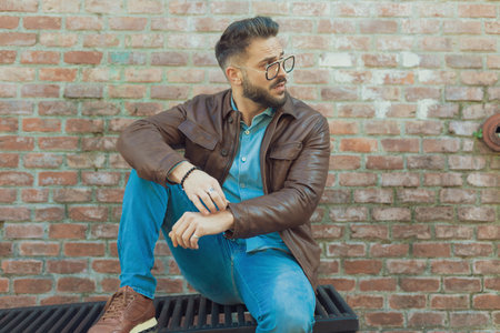 Portrait of handsome casual man sitting on a grill and looking away, wearing eyeglasses and leather jacket, outdoor in an old medieval townの写真素材