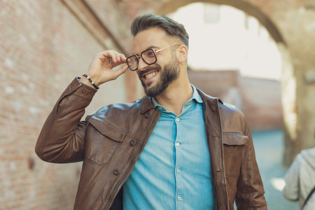 Portrait of attractive casual man arranging his glasses and laughing, wearing leather jacket, outdoor in an old medieval townの写真素材