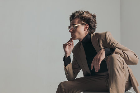 side view of fashion young man with blonde hair holding elbow on knee and thinking while looking to side on grey backgroundの写真素材