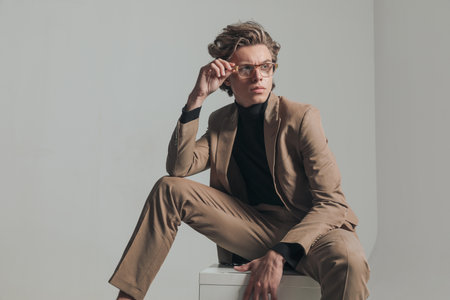 smart casual man sitting and holding elbow on knee while fixing glasses and looking to side on grey background in studioの写真素材