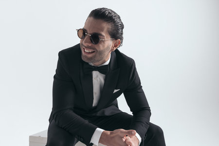 happy young man with sunglasses in black tuxedo looking to side and laughing while sitting with elbows on knees on grey backgroundの写真素材
