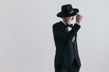sexy fashion guy in black tuxedo covering forehead and eyes with hat and posing in a confident way in front of grey backgroundの写真素材