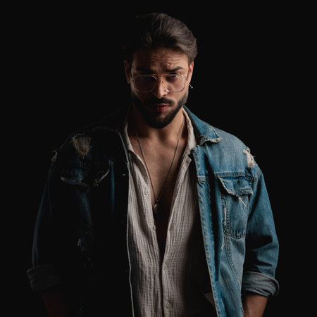 cool sexy fuy in denim jacket looking over glasses and posing in front of black background
