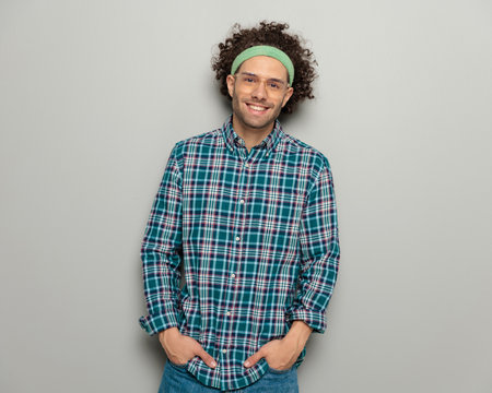 casual young man with glasses wewaring sport headband and smiling while posing with hands in pockets on grey backgroundの写真素材