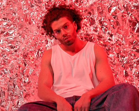 cool man with curly hair in t-shirt and jeans posing in a confident way with elbows on thighs in front of silver tinfoil background with red lightの写真素材