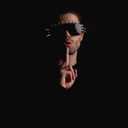 sexy mysterious guy with cool glasses making silence gesture and posing in a confident way in front of black backgroundの写真素材