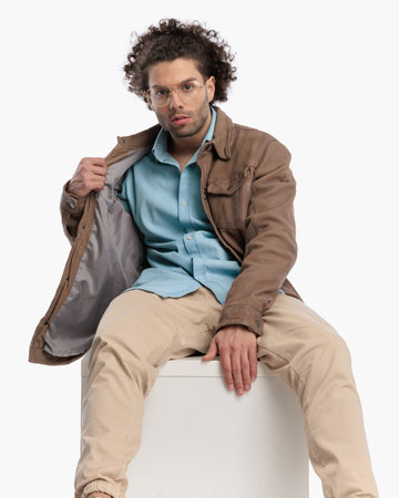 sexy man with curly hair and glasses adjusting beige jacket and sitting in front of white background in studioの写真素材