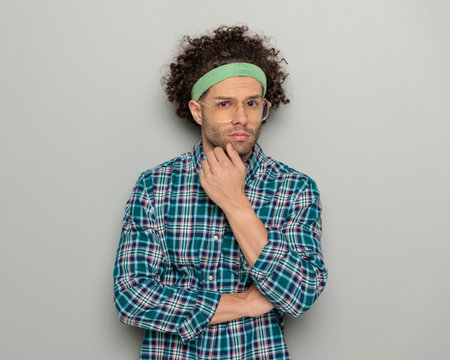sexy curly hair guy with sport headband and glasses touching chin and folding arms in front of grey backgroundの写真素材
