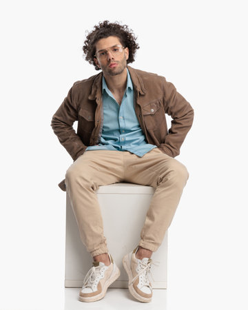 confident curly hair guy in beige jacket holding hands in pockets and posing in a cool way while sitting on white backgroundの写真素材