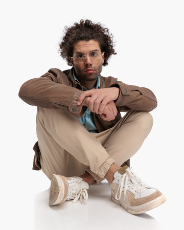 sexy curly hair man with glasses holding elbow on knee and sitting on the floor in front of white background in studioの写真素材