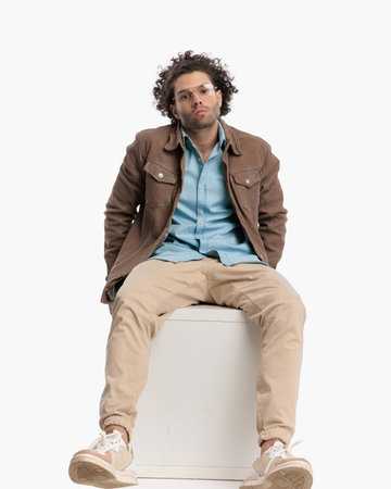 cool fashion guy with glasses and curly hair posing while sitting in a confident way in front of white background in studioの写真素材