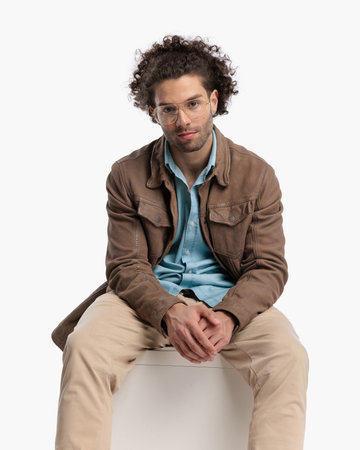 attractive casual man with glasses and curly hair sitting with arms on thighs in front of white background in studioの写真素材