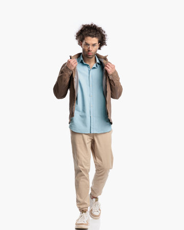 full body picture of cool casual guy walking and adjusting jacket in front of white background in studioの写真素材