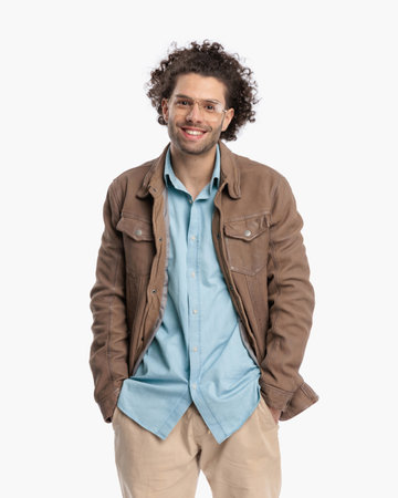 happy casual guy with glasses and curly hair holding hands in pockets and smiling in front of white background in studioの写真素材