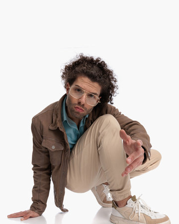 crouched cool young man with glasses showing hand forward and posing in front of white background in studioの写真素材