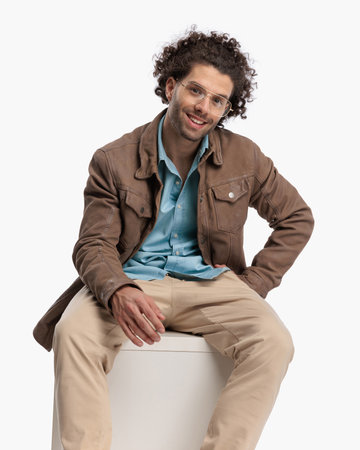 handsome young man with glasses wearing beige jacket, holding hand in pocket and smiling while sitting in front of white background in studioの写真素材