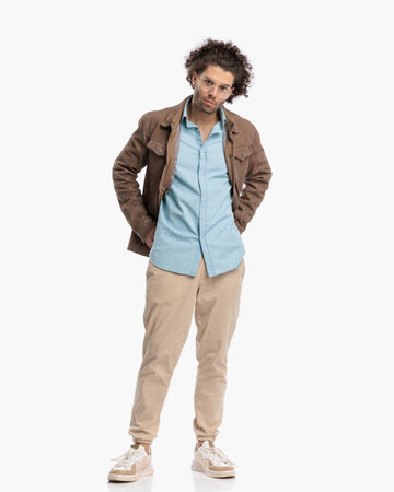 young casul man with curly hair holding hands in back pockets and posing while standing on white backgroundの写真素材