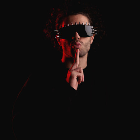mysterious young man wearing cool sunglasses and making silence gesture while hiding from police in front of black background with red lights onの写真素材