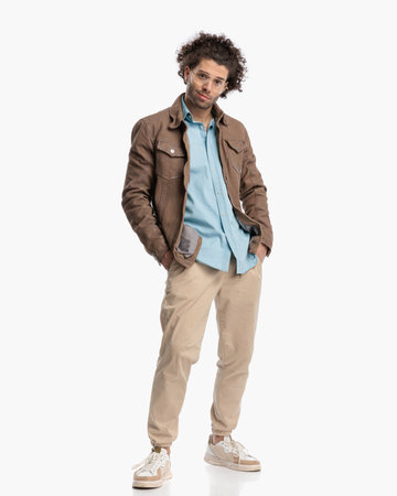 sexy happy guy with glasses and curly hair wearing beige jacket, holding hands in pockets and standing in front of white backgroundの写真素材