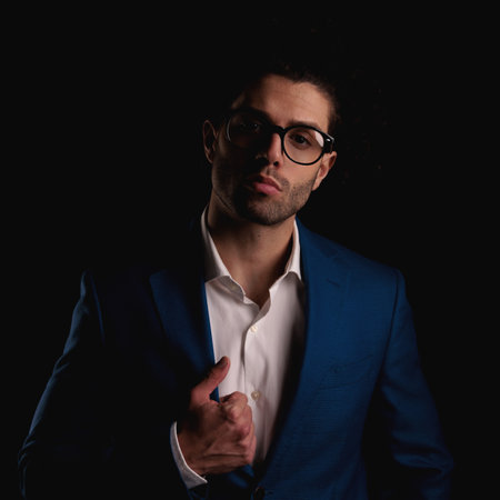attractive man with glasses and untied shirt holding and adjusting suit while confidently posing in front of black backgroundの写真素材