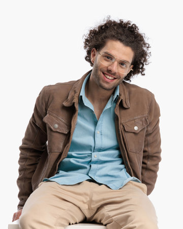 happy casual man with curly hair in beige jacket smiling and sitting in front of white background in studioの写真素材