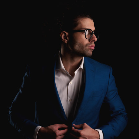 confident elegant man with glasses and open collar shirt buttoning suit while looking to side in front of black backgroundの写真素材