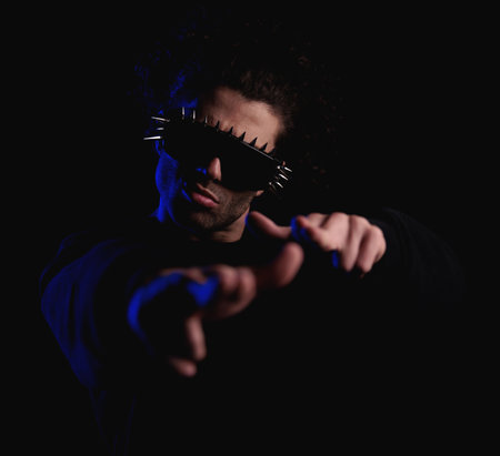 portrait of sexy man with curly hair wearing cool sunglasses and pointing hands forward in front of black background with blue lights onの写真素材